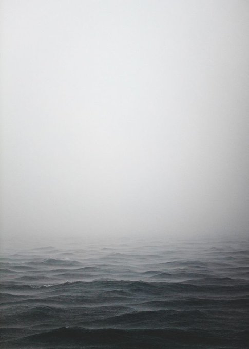 A moody seascape features dark, choppy ocean waves beneath a thick, gray, foggy sky.