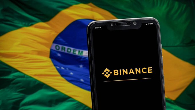 The Binance logo is displayed on a smartphone screen, set against the backdrop of the Brazilian flag. This image illustrates Binance's significant reduction of services and token suspensions affecting Brazilian users.