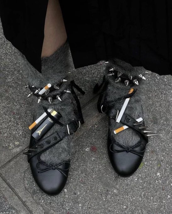A pair of black pointed-toe shoes featuring straps, metallic spikes, and what appear to be cigarettes tucked into the design, worn with fuzzy gray socks on a stone surface.