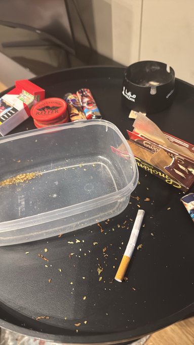 A messy black table containing an empty clear plastic container, a crushed cigarette, and an ashtray, scattered with tobacco and various snack wrappers.