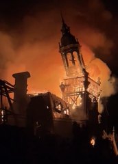 A large church or cathedral with a tall spire is engulfed in bright orange flames against the night sky.