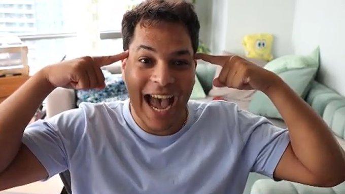 A young man in a light blue t-shirt sits indoors with a wide smile, pointing both index fingers at his temples in an animated, excited gesture.