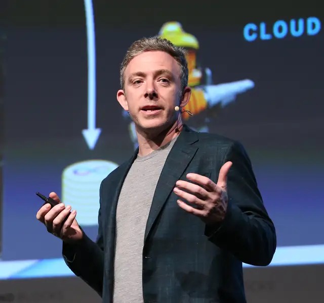 A man in a dark blazer and grey t-shirt speaking on stage while gesturing with his hands, holding a clicker, with a presentation screen behind him displaying the word 'CLOUD' and an arrow.