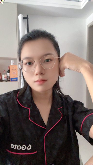 A young woman wearing glasses and black pajamas with pink trim poses for a selfie, with the mirrored text "Gate" visible on her top. The image illustrates the post's caption about receiving a GateQixi limited gift box featuring soft pajamas for relaxing at home after trading market ups and downs.