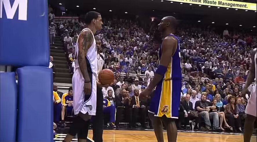 The image shows two basketball players standing next to each other on a court with one player holding a basketball while the other has his arms crossed over his chest, possibly greeting or discussing something with him. The scene takes place during a game, as evidenced by the presence of chairs and people in the background who are watching the match.