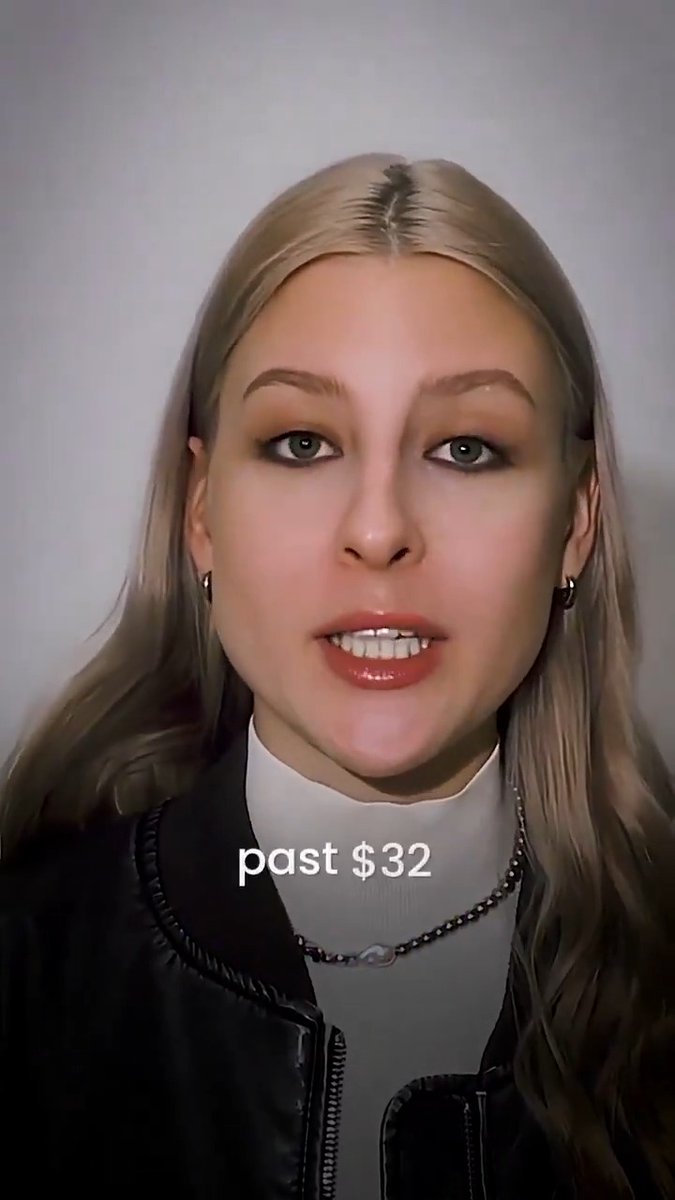A close-up selfie-style video of a blonde woman wearing a black jacket and white top, with on-screen text reading 'past $32'.