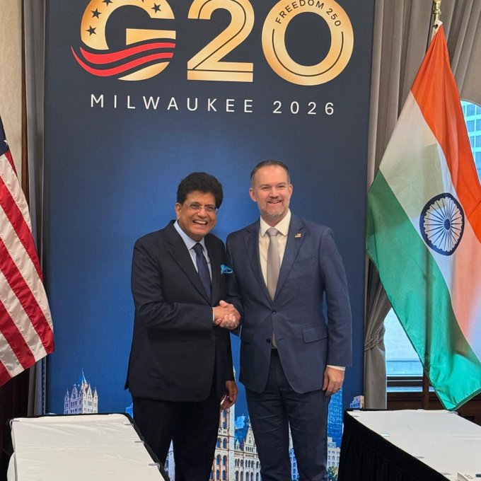 The image shows two men shaking hands and smiling in front of a backdrop reading "G20 MILWAUKEE 2026" and "FREEDOM 250," flanked by the American and Indian flags. The man on the left appears to be Piyush Goyal and the man on the right USTR Jamieson Greer, visually capturing the meeting referenced in the post about advancing the India-U.S. interim trade deal.