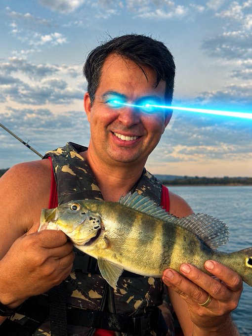 A smiling man in a camouflage life vest holds up a freshly caught perch on a lake at sunset, with digitally added glowing blue laser beams shooting from his eyes.