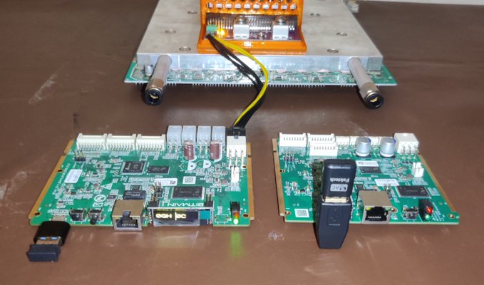 Two green printed circuit boards (PCBs) sit on a wooden surface, connected by yellow and black wires to a larger silver heatsink assembly above them, featuring various ports, capacitors, and chips.