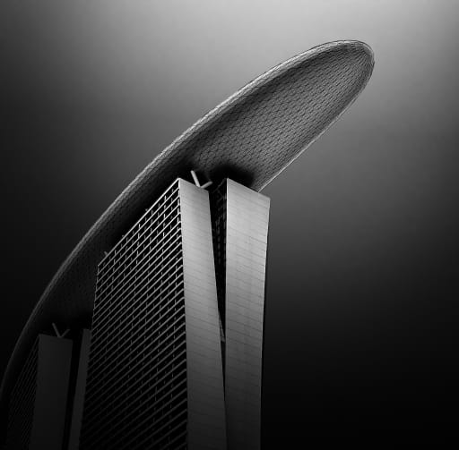 A dramatic black and white photograph of the iconic Marina Bay Sands hotel in Singapore, showing its three towers topped by the boat-shaped SkyPark against a dark sky.