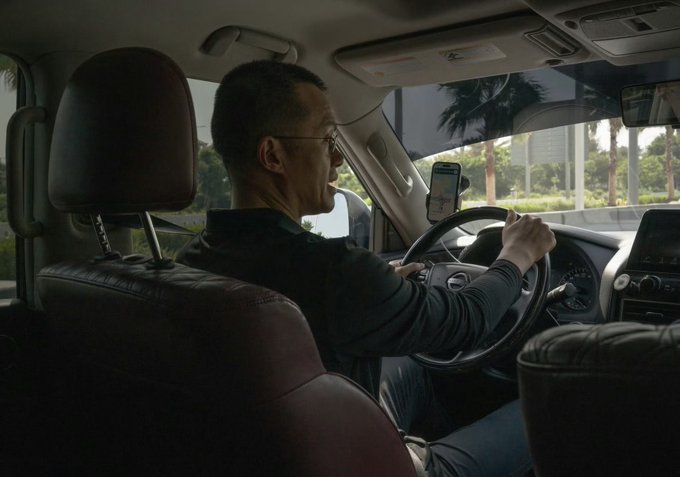 A candid photograph of an older man wearing glasses driving a vehicle, with a smartphone mounted on the dashboard displaying navigation, captured in a realistic, photojournalistic manner.