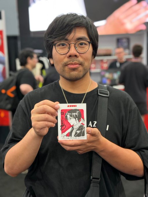 A man with dark hair and round glasses stands at what appears to be a convention, proudly displaying a small manga print labeled "AZUKI" featuring a character illustration of himself. The caption celebrates his collaboration with Azuki and Steamboy33, where the print represents his personal contribution to the manga IP that he and his partner have been involved with for nearly five years.