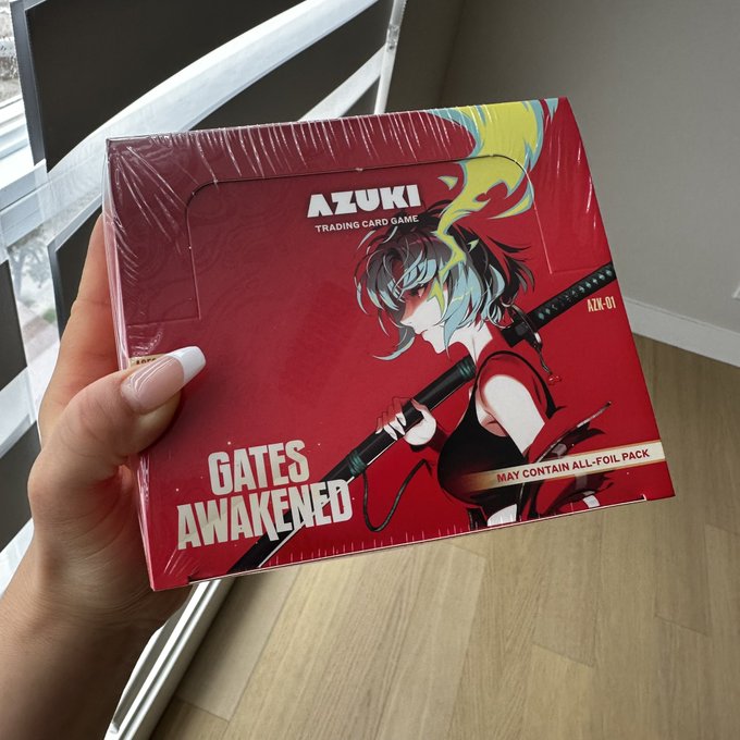 A hand holds a sealed "Azuki Trading Card Game - Gates Awakened" box (AZK-01), featuring a red-haired anime character with flame motifs, capturing the unopened collectible referenced in the post.