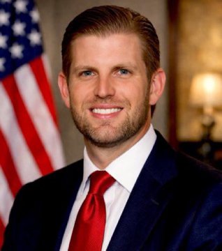 Official portrait of Eric Trump wearing a dark suit with a red tie, posed in front of an American flag.