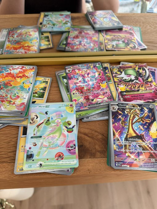 A collection of Pokémon trading cards, including Mew VMAX, Gengar VMAX, Starmie V, and Charizard, is spread across a wooden table alongside several stacked Elite Trainer Boxes (ETBs), showcasing an impressive haul of booster packs and valuable cards.