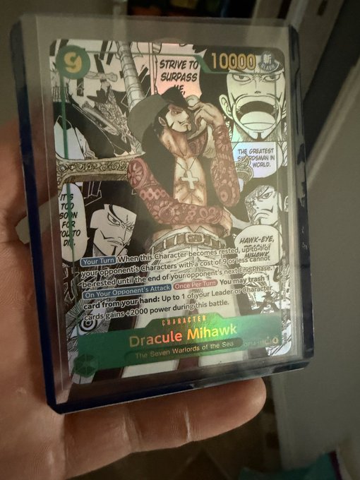A person holding a holographic One Piece trading card featuring the character Dracule Mihawk, which has a power of 10000 and detailed game text for its abilities.