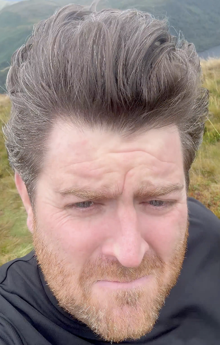 A close-up selfie shows a bearded man with tousled hair squinting in a pained or stressed expression outdoors, with grassy hills and water visible in the background.