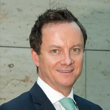 A professional headshot of a man in a dark suit and light green tie, identified by context as Standard Chartered's Geoff Kendrick, whose Bitcoin price forecast has been revised upward.