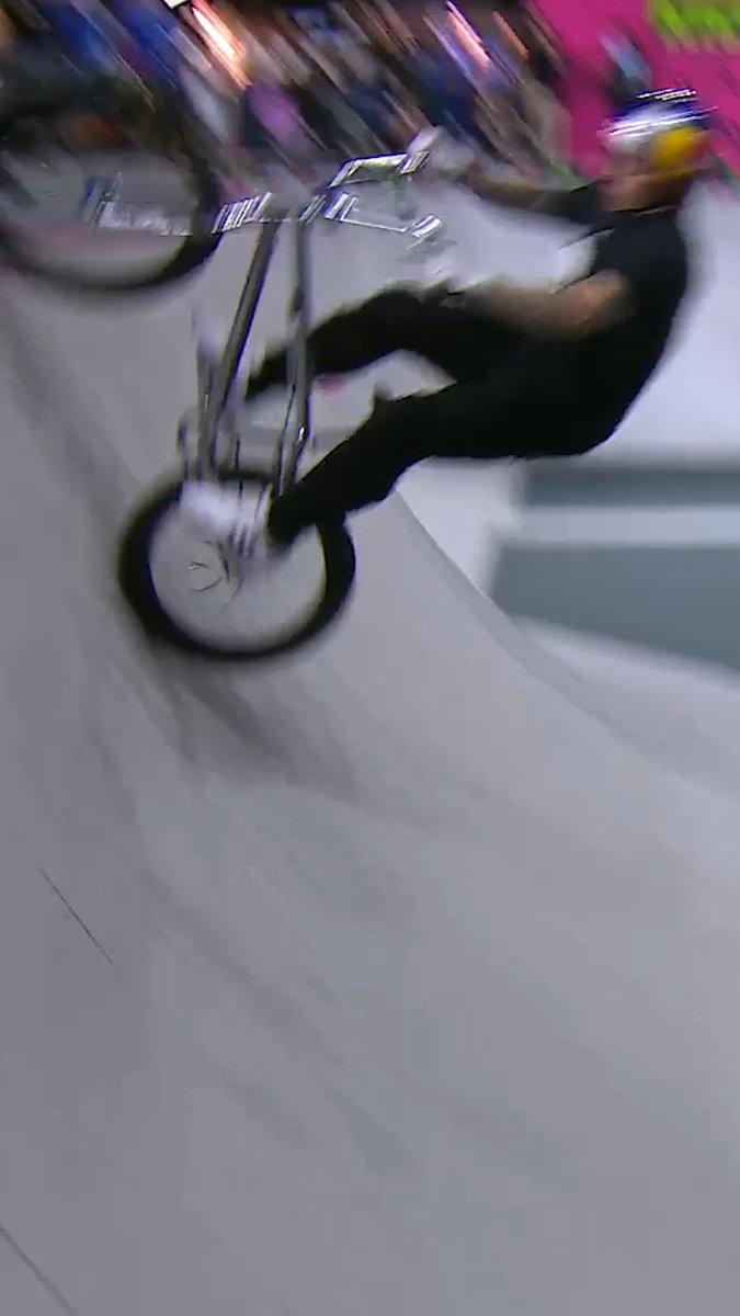 A BMX rider performs an aerial trick on a ramp in a skatepark, wearing a helmet and dark clothing, with motion blur capturing the fast-paced action of the competition. The image aligns with the caption describing Kieran Reilly's bronze-winning flair double tailwhip to barspin at the Dave Mirra BMX Park Best Trick event during the X Games League Championship in New Orleans.