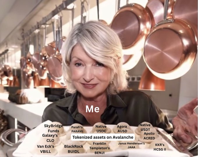The image features a woman with blonde hair standing next to a table filled with various pots, pans, and other kitchen items such as spoons and bowls. The woman appears to be smiling at the camera while holding up a plate of food or possibly serving herself from the dish on the table. In the background, there are several pots hanging above her head, adding to the overall atmosphere of a busy kitchen scene.