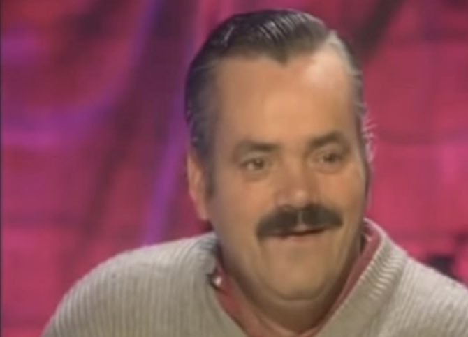 A man with dark hair and a thick mustache, wearing a light-colored sweater over a collared shirt, smiles against a pink and purple studio background.
