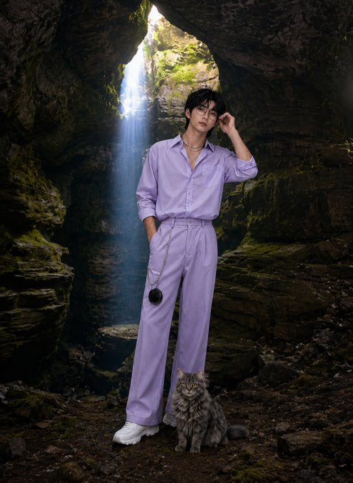 A young person in a lavender shirt and matching wide-leg trousers poses in a mossy cave with a waterfall behind them, accompanied by a fluffy gray tabby cat at their feet. The ethereal, mystical setting complements the "GM CT" greeting, evoking a serene, otherworldly "good morning" atmosphere.