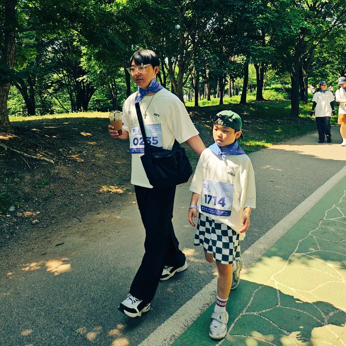 The image shows a man and a child walking down a path, both wearing shirts with numbers on them. They are accompanied by other people who appear to be participating in an event or race. The scene suggests that they might be taking part in a cycling event or a similar outdoor activity.