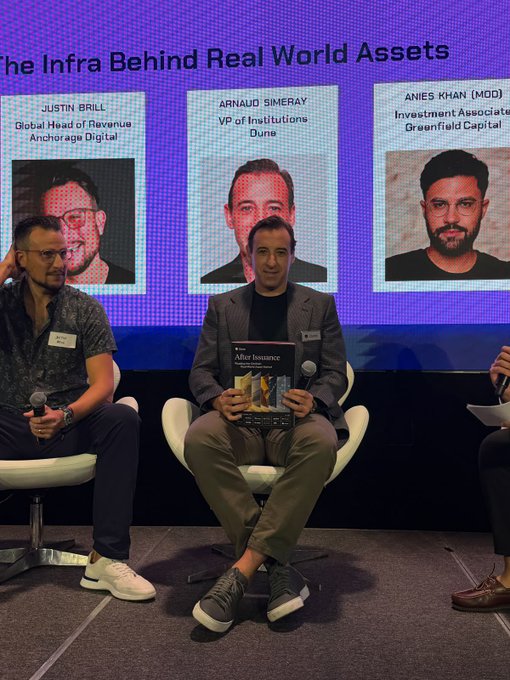 Two men sit on stage during a panel titled "The Infra Behind Real World Assets," with one holding up an "After Issuance" magazine/booklet, perfectly executing the "RWA bible has arrived" assignment as featured panelists Justin Brill (Anchorage Digital), Arnaud Simeray (Dune), and moderator Anies Khan (Greenfield Capital) are highlighted on the screen behind them.