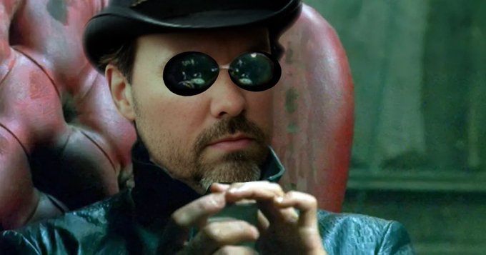 A close-up of a bearded man wearing a black top hat, round dark sunglasses, and a dark leather coat, holding a small object while seated against a tufted red leather chair.