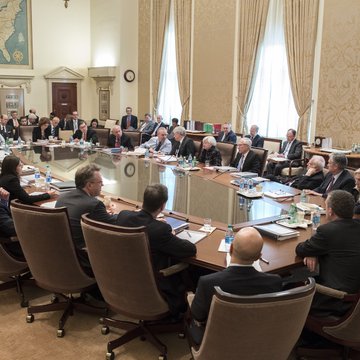 The image shows a group of people sitting around a conference table, engaged in a meeting or discussion. The room has large windows that allow natural light into the space, creating an atmosphere conducive to conversation. There are several chairs placed around the table, accommodating the attendees. A clock is visible on one wall, allowing participants to keep track of time during their gathering.

The people present appear to be professionals, possibly involved in financial or business matters. The room has a formal setting with a large window and a brown carpeted floor. There are multiple books scattered around the table, suggesting that they may be discussing important topics related to finance or other subjects. A bottle is also visible on one side of the table, indicating refreshments might have been provided for the attendees during their meeting.