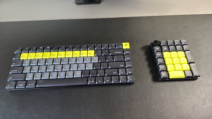 Two black and yellow mechanical keyboard components rest on a dark desk mat, including a main alphanumeric keyboard and a separate macro pad.