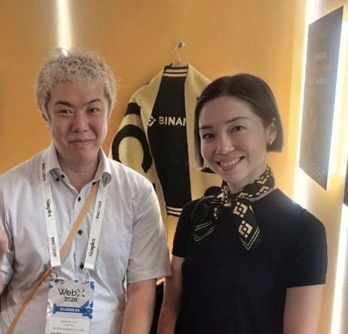 A man wearing a "WebX 2026" lanyard badge poses for a photo alongside a woman, with a yellow Binance-branded jacket hanging on the wall behind them, commemorating their meeting at the WebX 2026 conference featuring a Binance Japan representative.