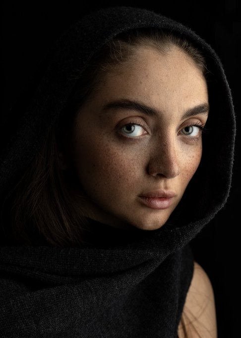 A close-up studio portrait of a woman wearing a black hooded wrap against a black background, with freckles and pale eyes clearly visible.