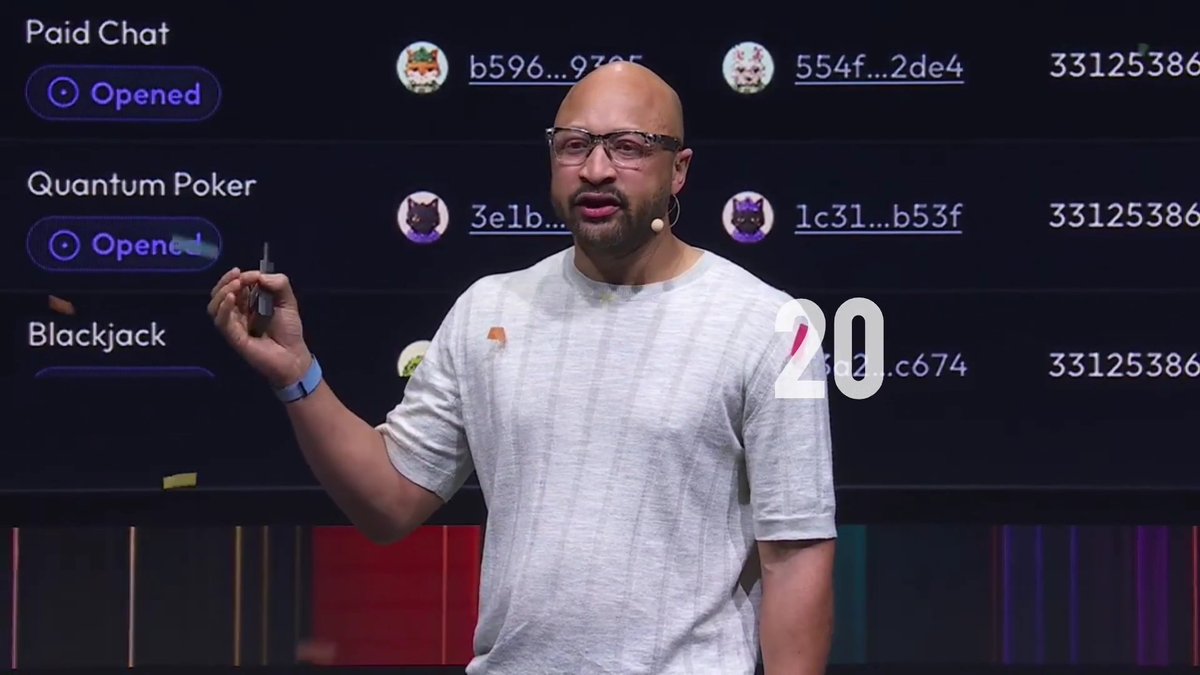 A bald man with glasses and a beard wearing a gray t-shirt giving a presentation on stage, with a screen behind him showing options like 'Paid Chat', 'Quantum Poker', and 'Blackjack' alongside crypto addresses.