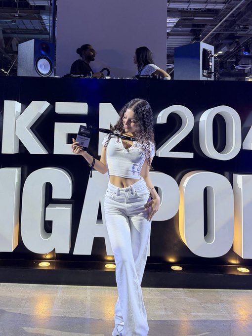 A young woman in a white crop top and white flare pants holding a microphone, standing in front of a large sign displaying 'K-... 20...' and 'GA... PO...' letters with DJs and speakers visible in the background.