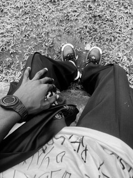 A black and white, first-person perspective photo looking down at a person's legs, sneakers, and a hand wearing a watch while sitting on grass scattered with leaves.