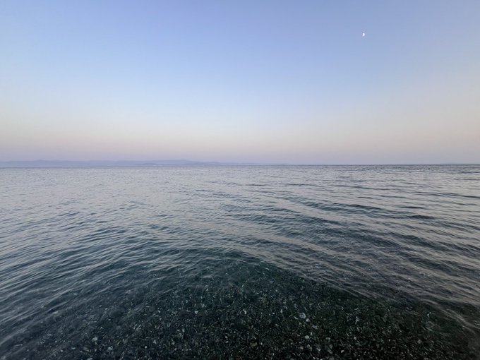 Serene photograph of a calm body of water with a pebble-stone shoreline in the foreground, mountains visible in the distance, and a faint moon in a pastel twilight sky.
