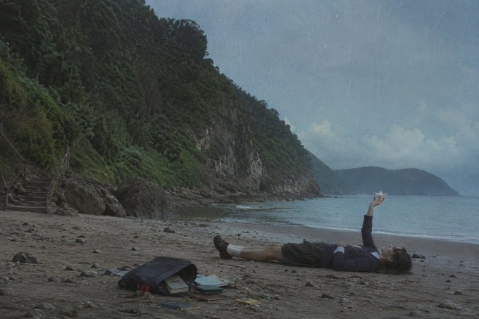 A moody, atmospheric photograph of a person lying on a rocky beach in the rain, holding up a small white origami star, with a dark, forested cliff and a bag of belongings nearby.