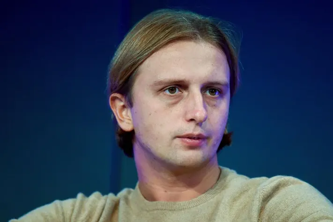 A caption describing the image: A man with medium-length blond hair and blue eyes, wearing a beige sweater, pictured against a dark blue background. The image likely relates to the Revolut article, possibly showing the fintech company's founder or a company representative, though no name or identifying text is visible in the image itself.