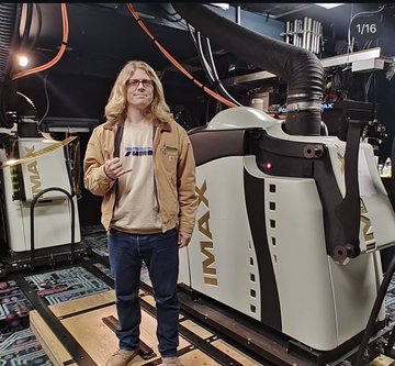 A blonde man wearing sunglasses and a tan jacket standing next to a large Maxx industrial machine in a warehouse/workshop setting.