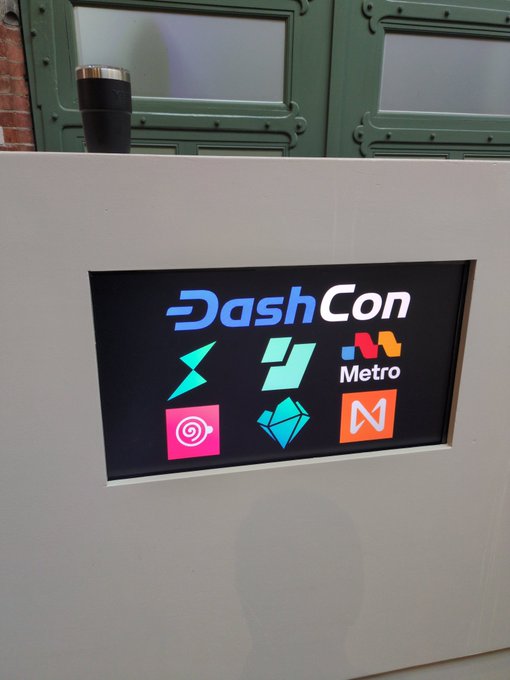 A black sign at what appears to be a convention displaying the 'DashCon' logo along with several cryptocurrency-related icons including Metro, Ethereum, and others.
