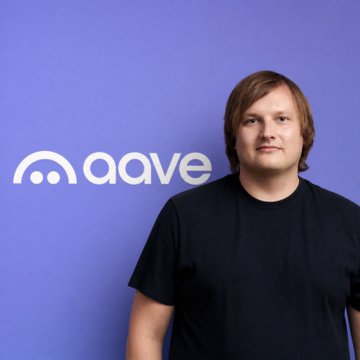 The image features a man standing against a purple background, wearing a black t-shirt with white text that says "Aave" written in capital letters. The man appears to be posing for a photo or portrait. There are no other objects or people visible in the image.