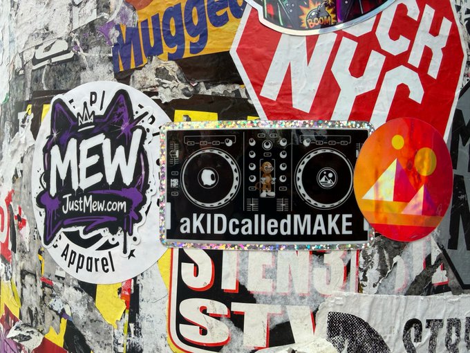 A collage of various stickers on a wall, including a NYK sticker, a circular 'MEW JustMew.com Apparel' sticker, a 'Mugged' sticker, and a central DJ deck sticker with text reading 'aKIDcalledMAKE'.