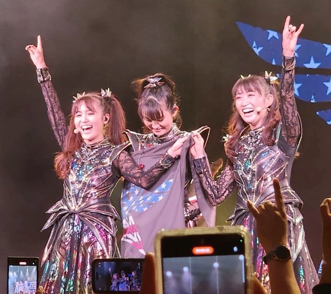 Three performers in elaborate sparkly costumes on stage with their arms raised, smiling, with audience members holding up smartphones in the foreground.