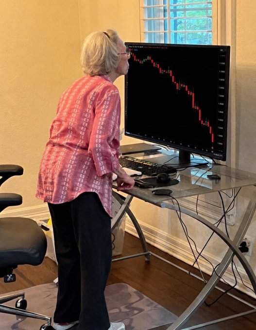 An elderly woman in a pink patterned shirt stands at a desk closely examining a computer monitor displaying a red downward-trending stock market candlestick chart.