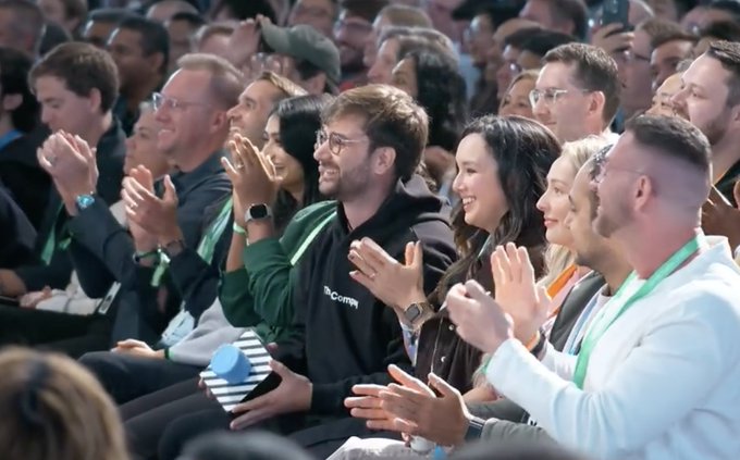 An audience applauding at an event, with attendees wearing lanyards and smiling as they clap.