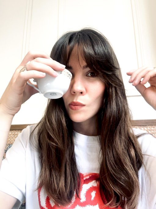 A young woman with long dark hair and bangs holds a small white teacup over her right eye, wearing a white t-shirt with a red graphic design.