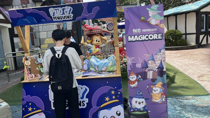 Two visitors pose in front of colorful promotional booths at Lotte World Adventure, with displays reading "Pudgy Penguins" and "MemeCore MAGICORE" featuring cartoon mascots and castle-themed artwork, reflecting the post about meeting the Pudgy Penguins and MemeCore brands at the park.