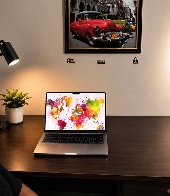 A desk setup with a laptop displaying a colorful world map wallpaper, a small potted plant, a black desk lamp, and a framed picture of classic cars hanging on a beige wall above small stickers.
