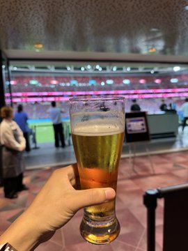 A first-person view of a hand holding a tall glass of beer in what appears to be an airport or large venue, with a check-in counter or service desk visible in the blurred background.